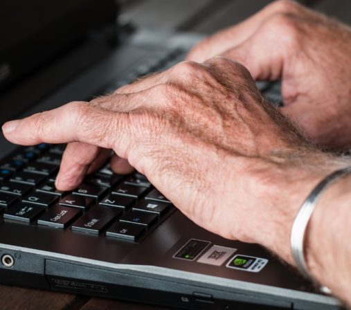 Tietokoneen näppäimillä näpyttelee vanhemman henkilön sormet. Etualalla olevan käden ranteessa on metallinen rannekoru.