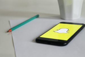 Keltain Snapchat-kuvake kännykän näytöllä. Vieressä vihreä lyijykynä ja paperia. 